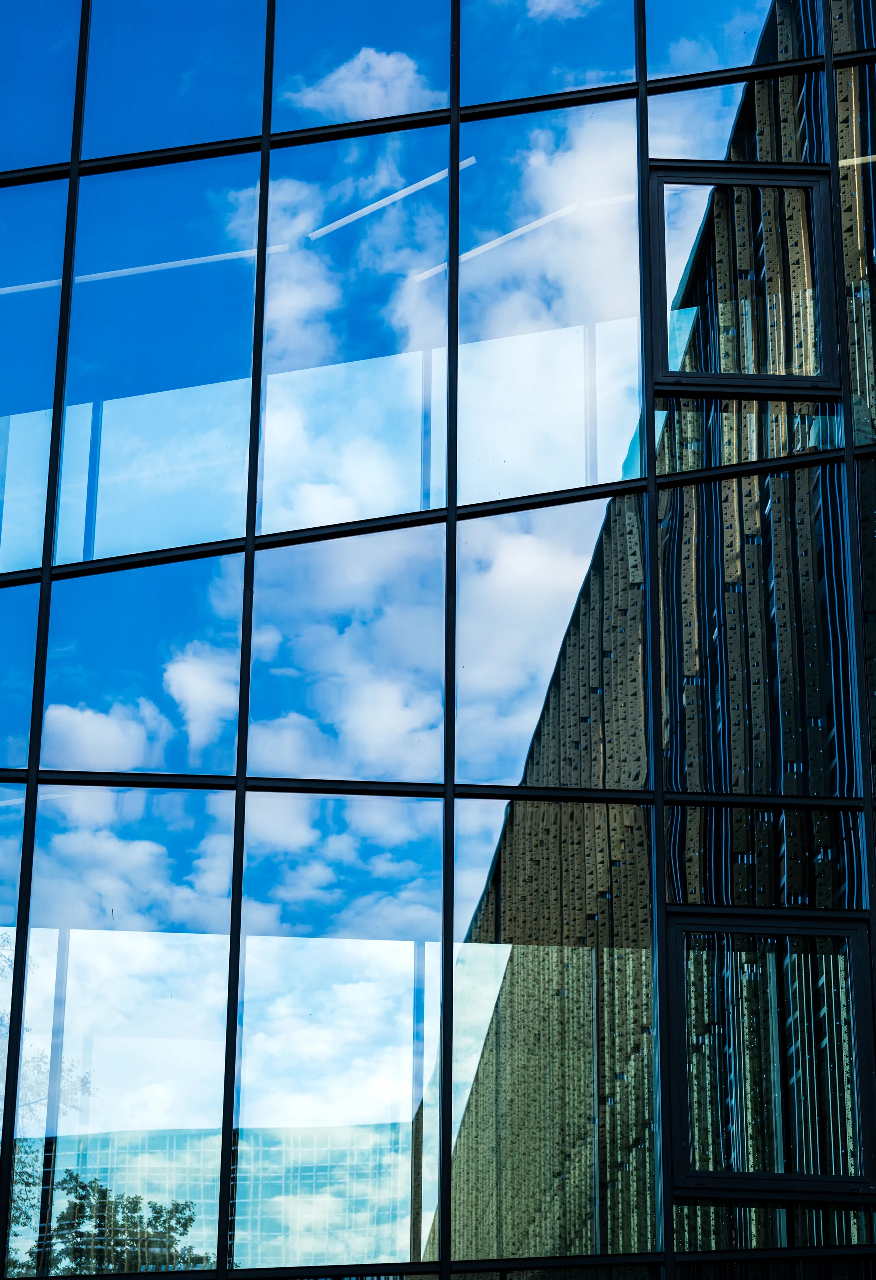 White clouds and blue sky reflected in the Delta Centre’s grid of glass panels, with golden facade fins on the right.