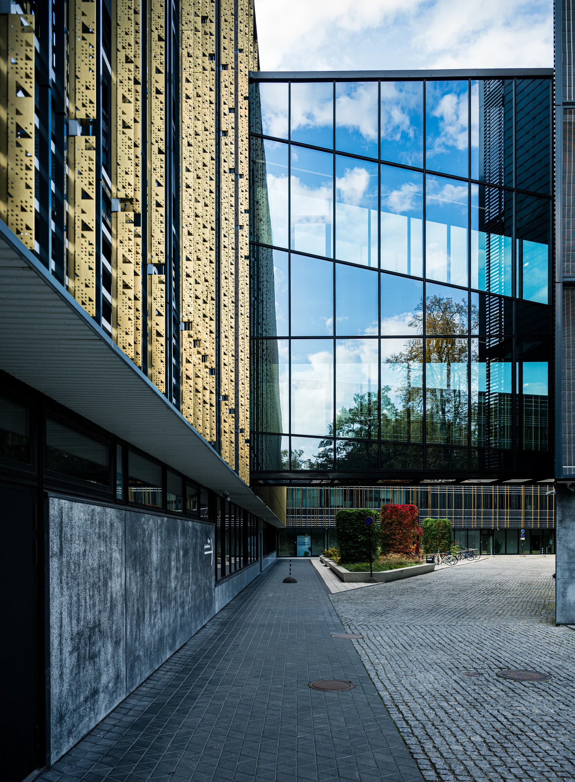 The paved courtyard of the Delta Centre beneath a glass connecting walkway, bordered by golden facade fins and grey walls.