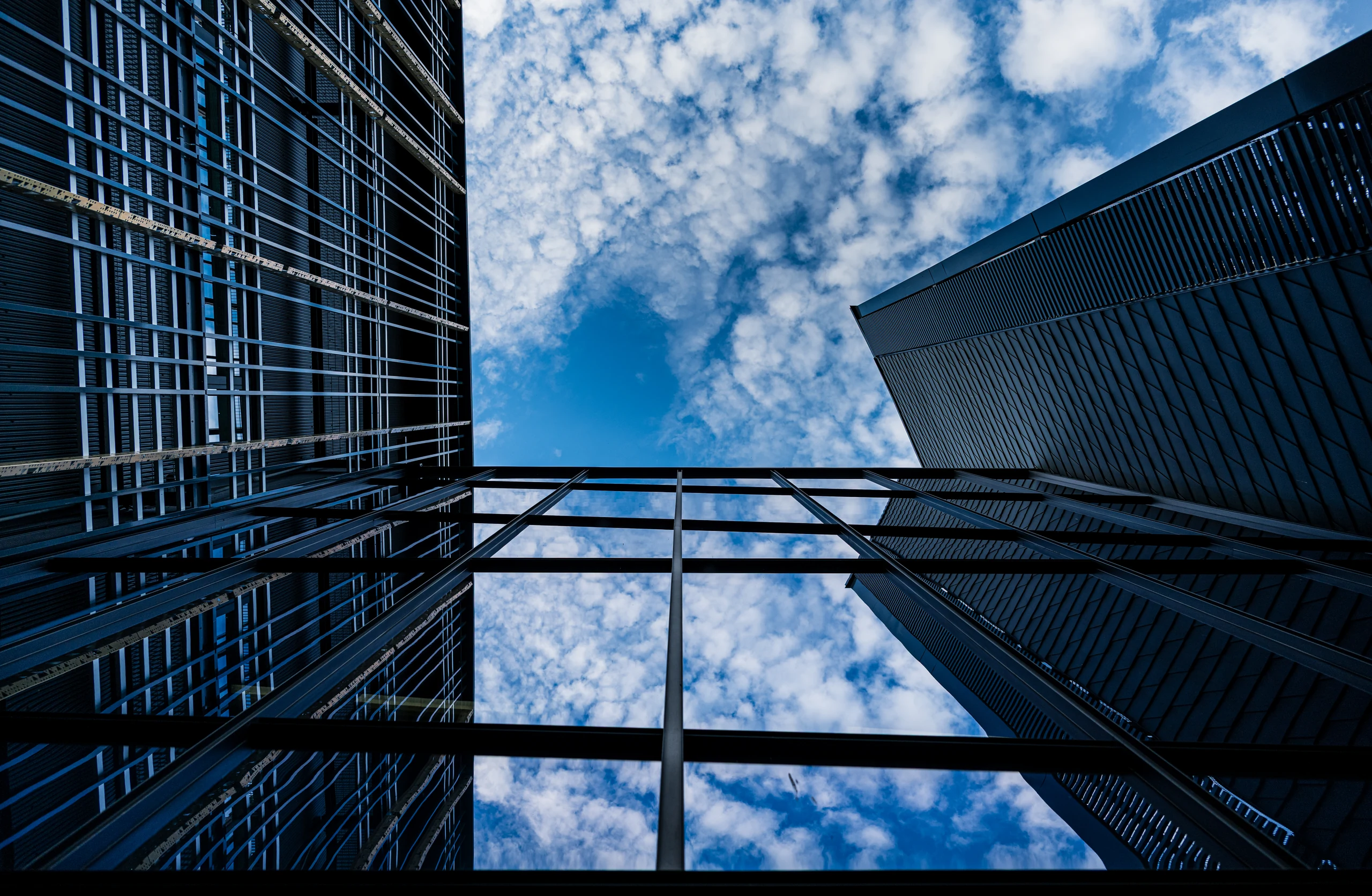 A view up between the Delta Centre’s buildings towards blue sky and clouds, reflected in the glass connecting walkway below.