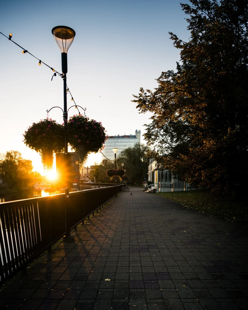 Morning sun shines onto the Emajõgi promenade, with a hanging flower basket on a lamp post and the riverside railing in the foreground.