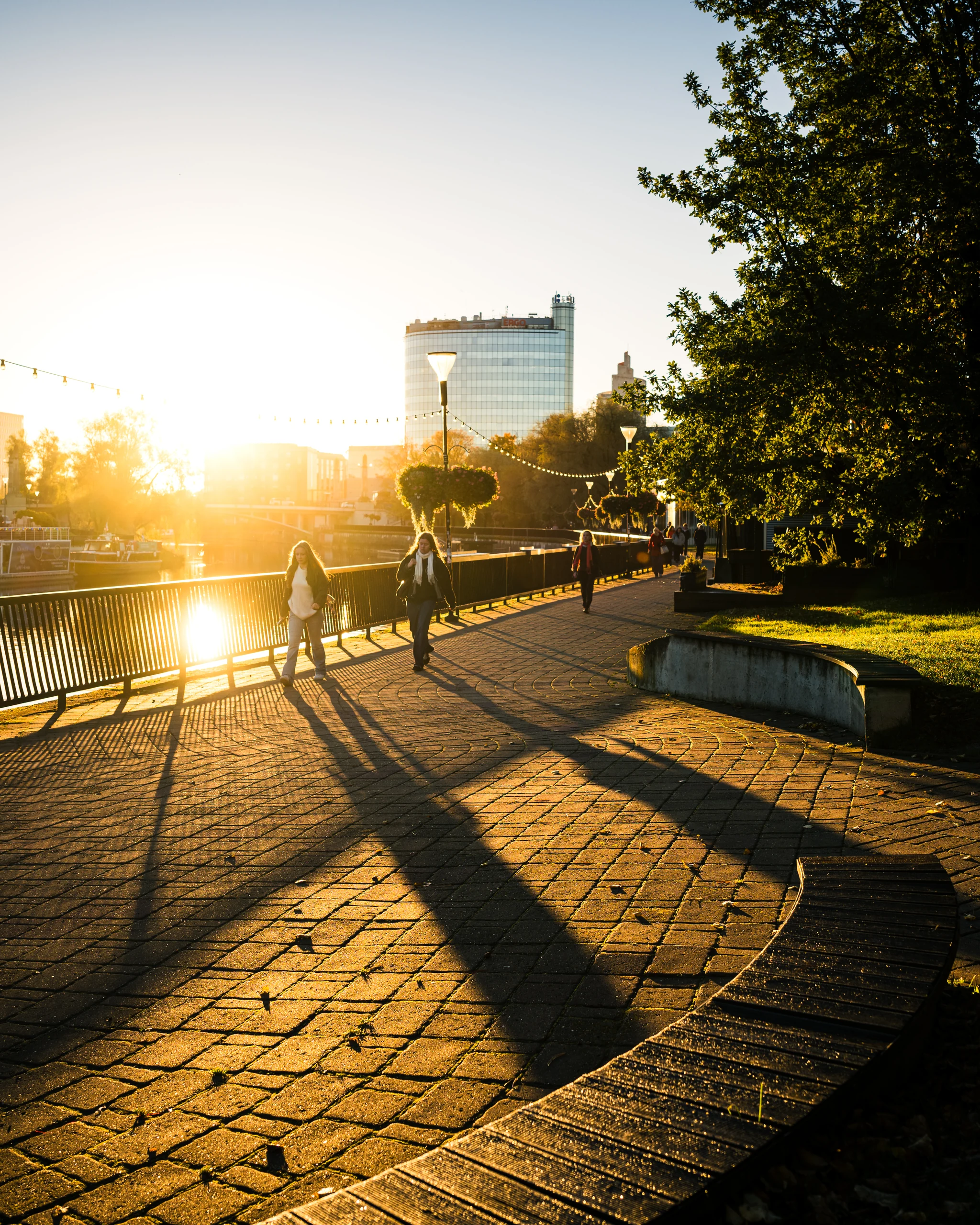 A pedestrian and a cyclist on the Emajõgi promenade, with morning sunlight casting long shadows across the paving.