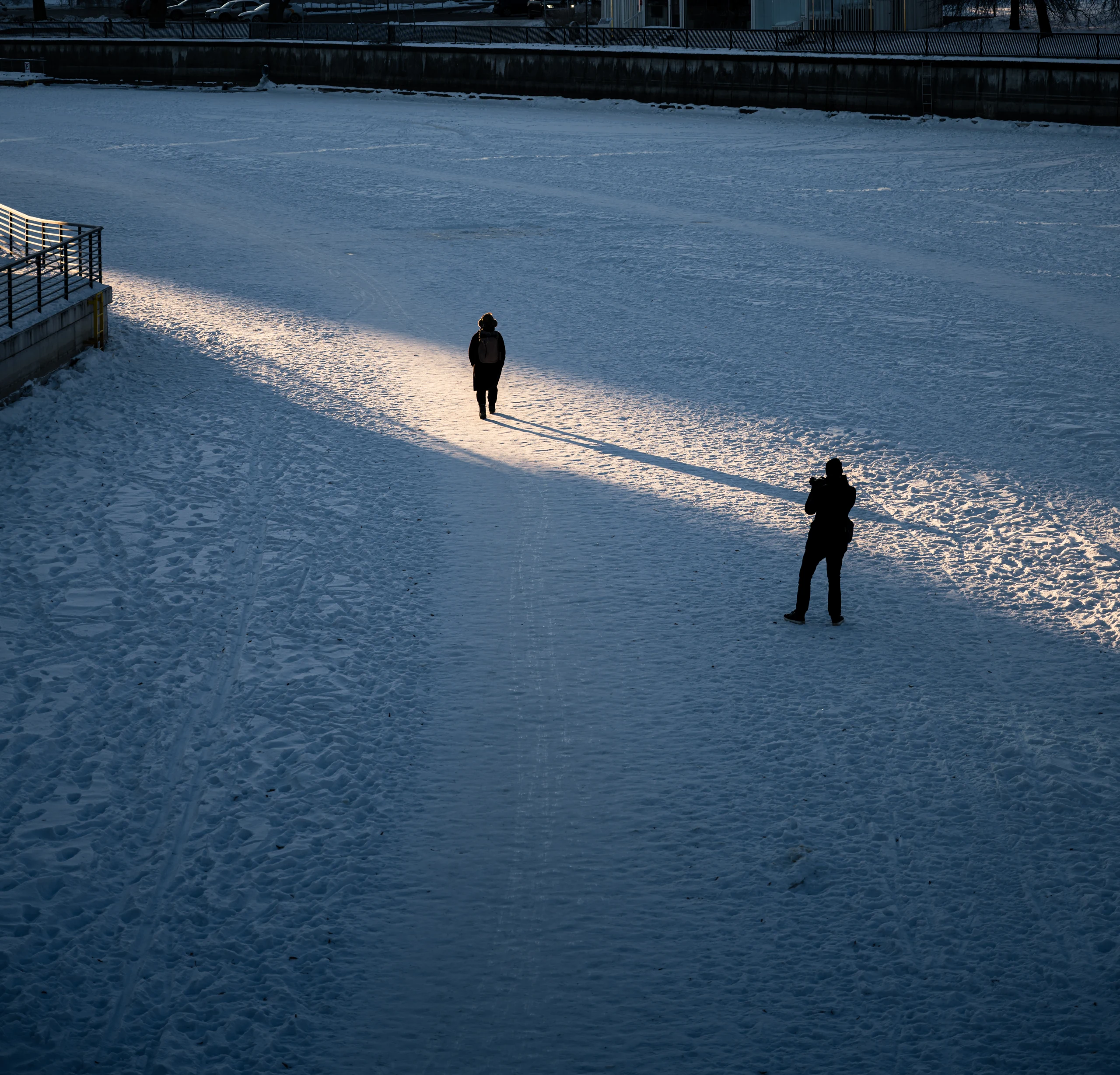 Two people on the snow-covered Emajõgi River, one in a strip of sunlight and the other in shadow.