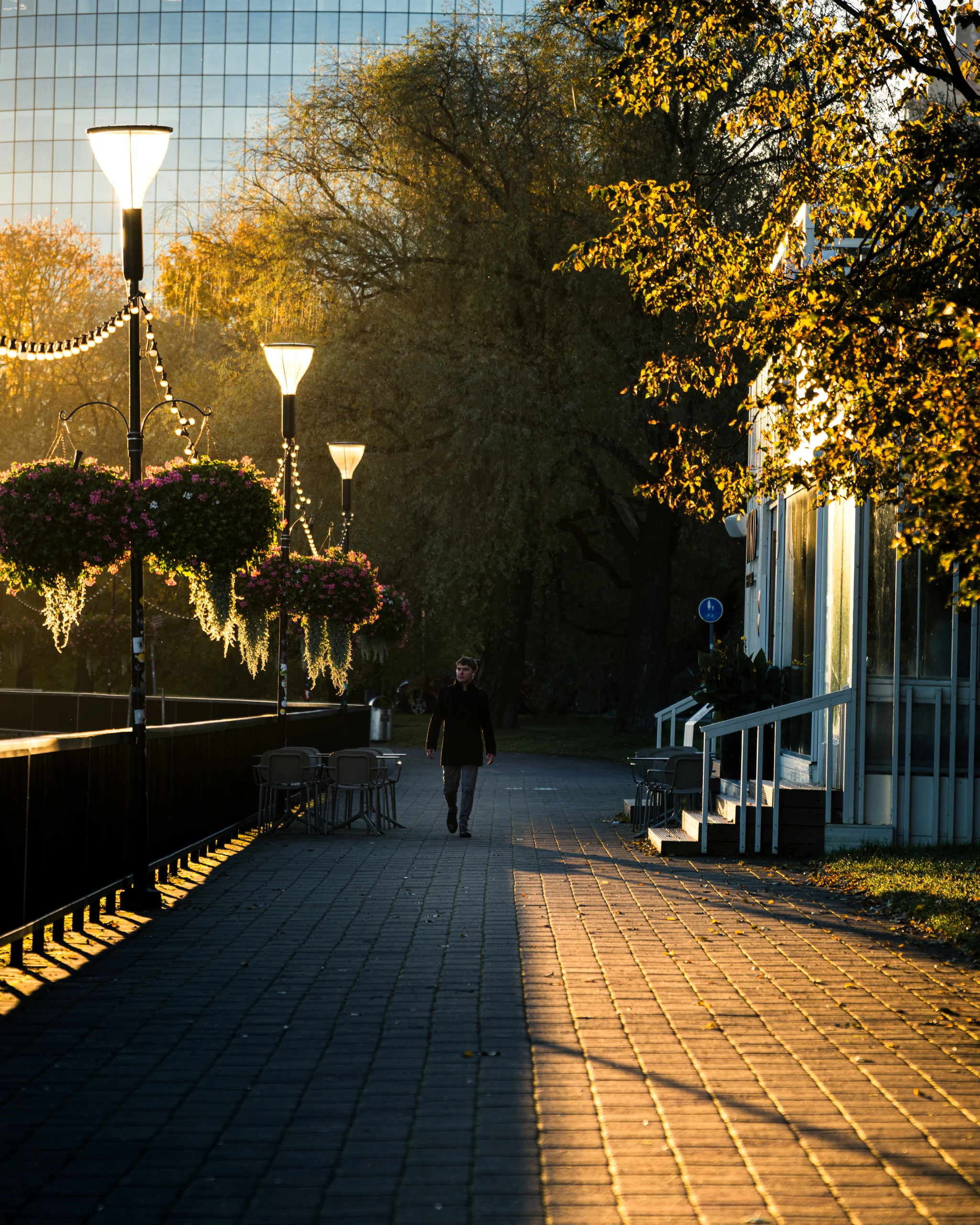 A person walks along the Emajõgi promenade between lamp posts, hanging flower baskets and autumn trees lit by golden sunlight.