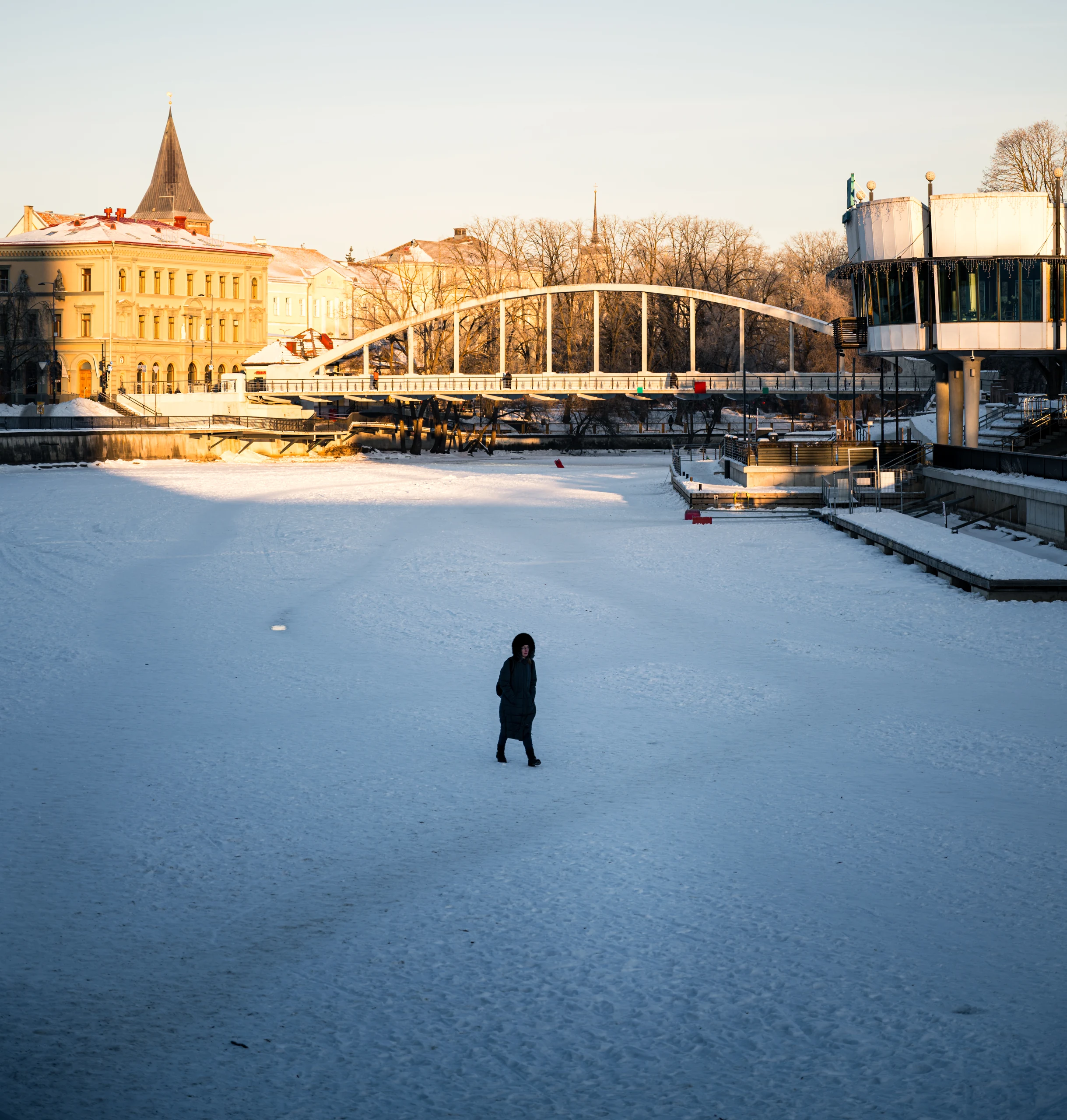 A lone person on the snow-covered Emajõgi River, with Kaarsild and sunlit city-centre buildings behind.