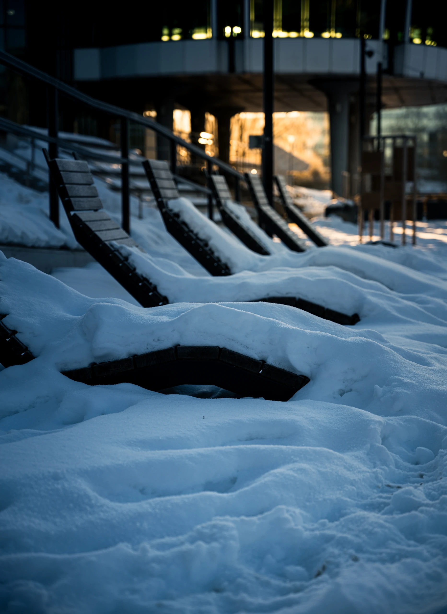 Curved lounge chairs covered in snow, with golden reflections in a glass facade behind them.