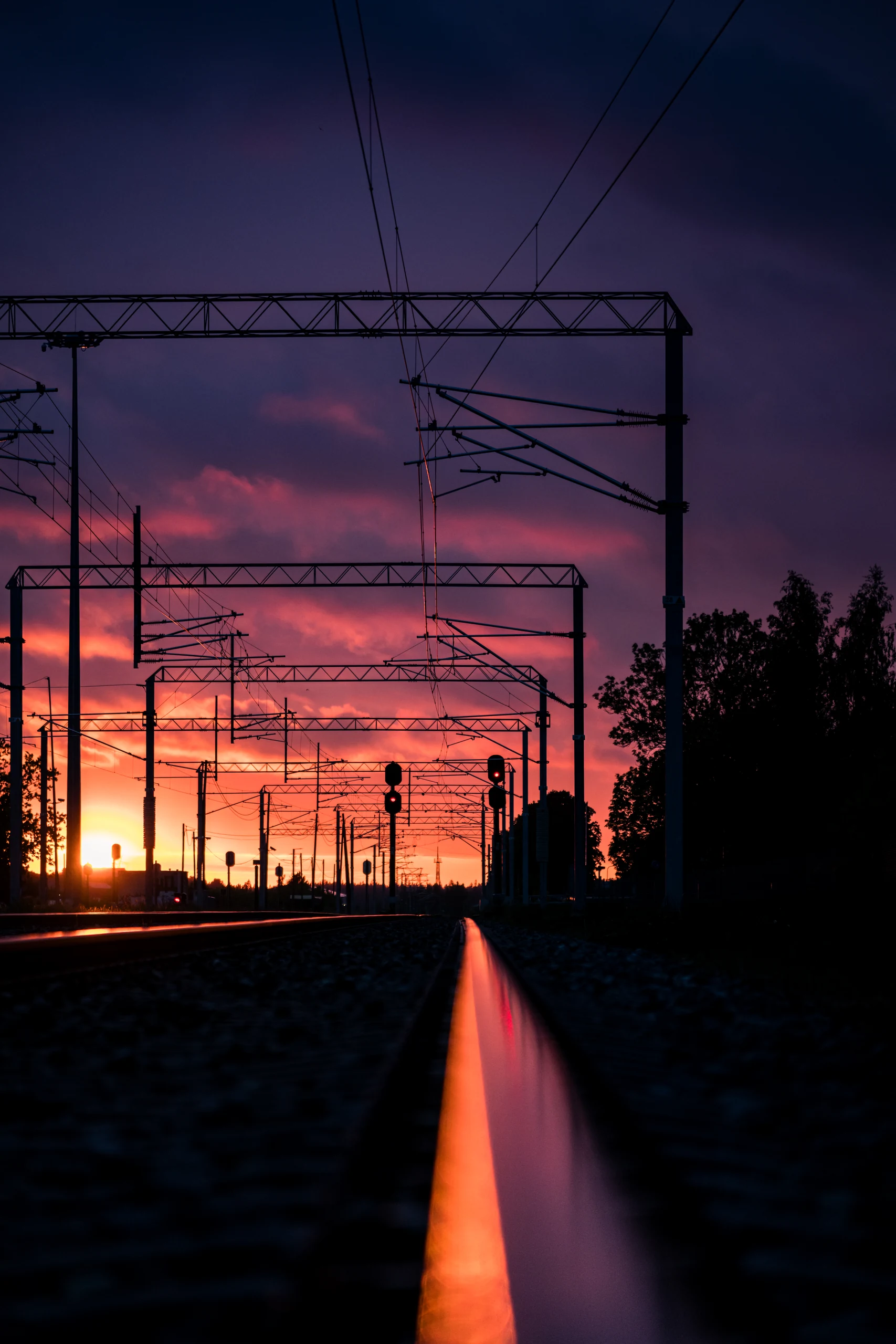 A rail glowing orange at sunset leads towards receding overhead line masts and red-purple clouds.