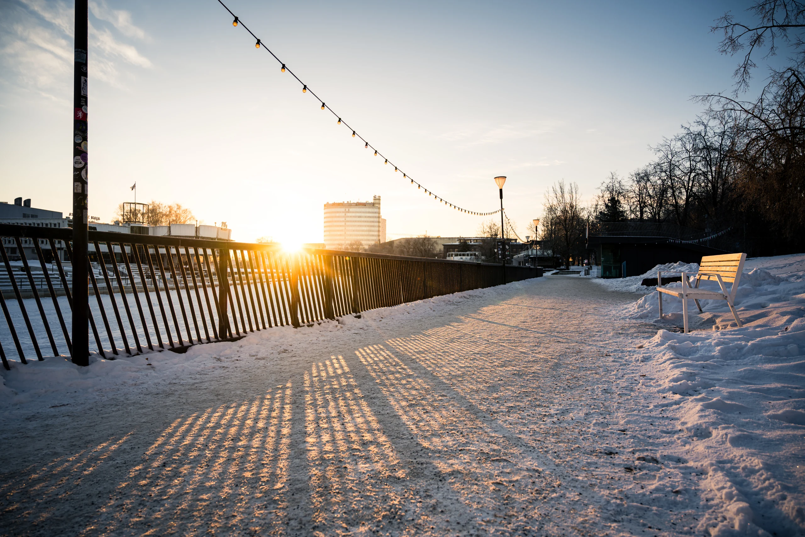 The snow-covered Emajõgi promenade, a white bench and long railing shadows in golden morning light.