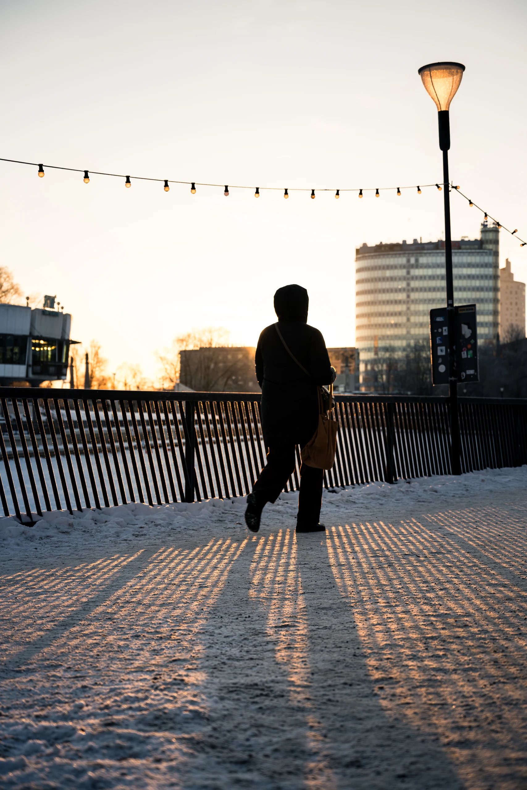 A walker in a dark coat on the snowy promenade, with railing shadows and a lamp in low sunlight.