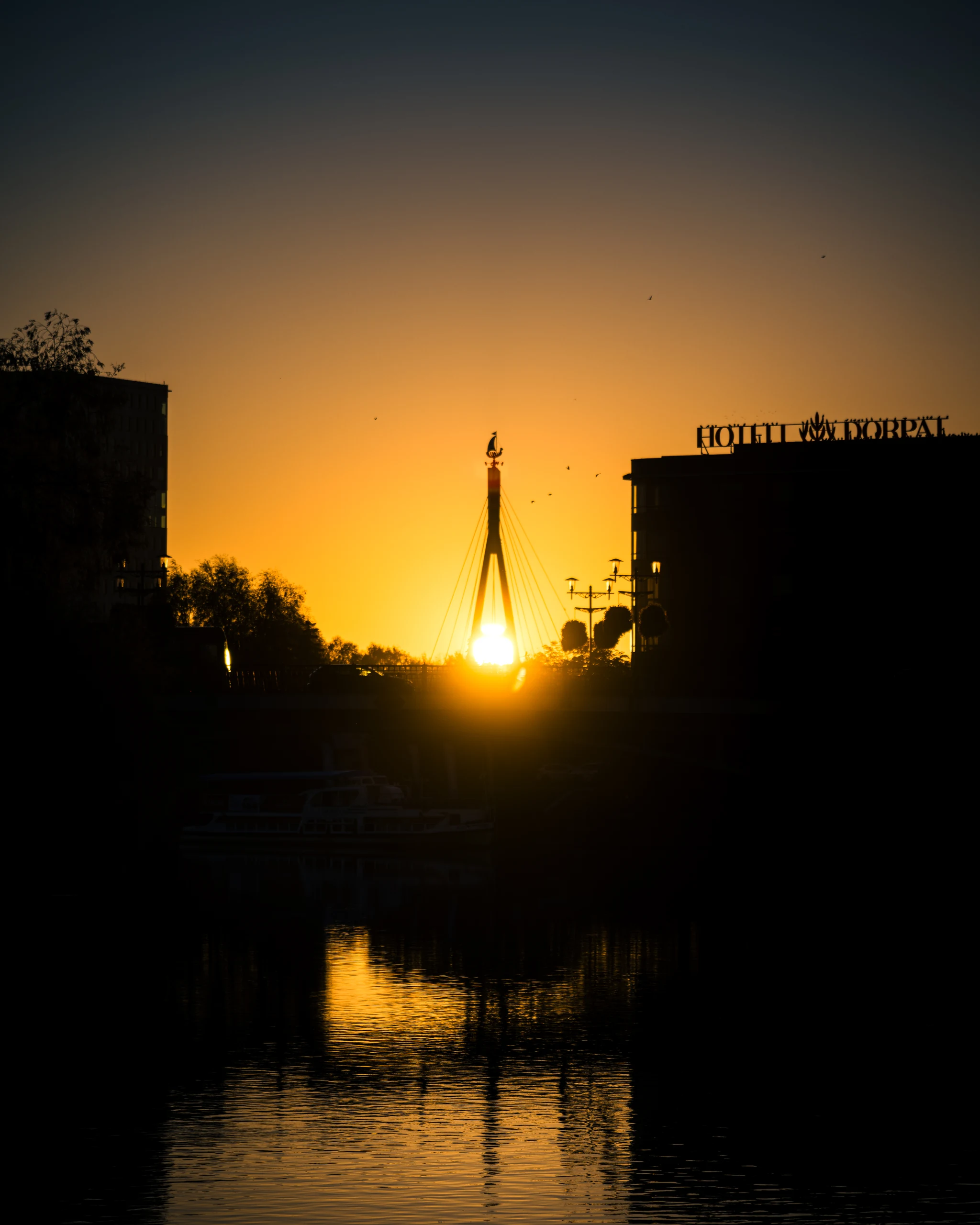 The rising sun beneath the pylon of Turusild, with a golden reflection on the Emajõgi River and dark silhouettes along the banks.