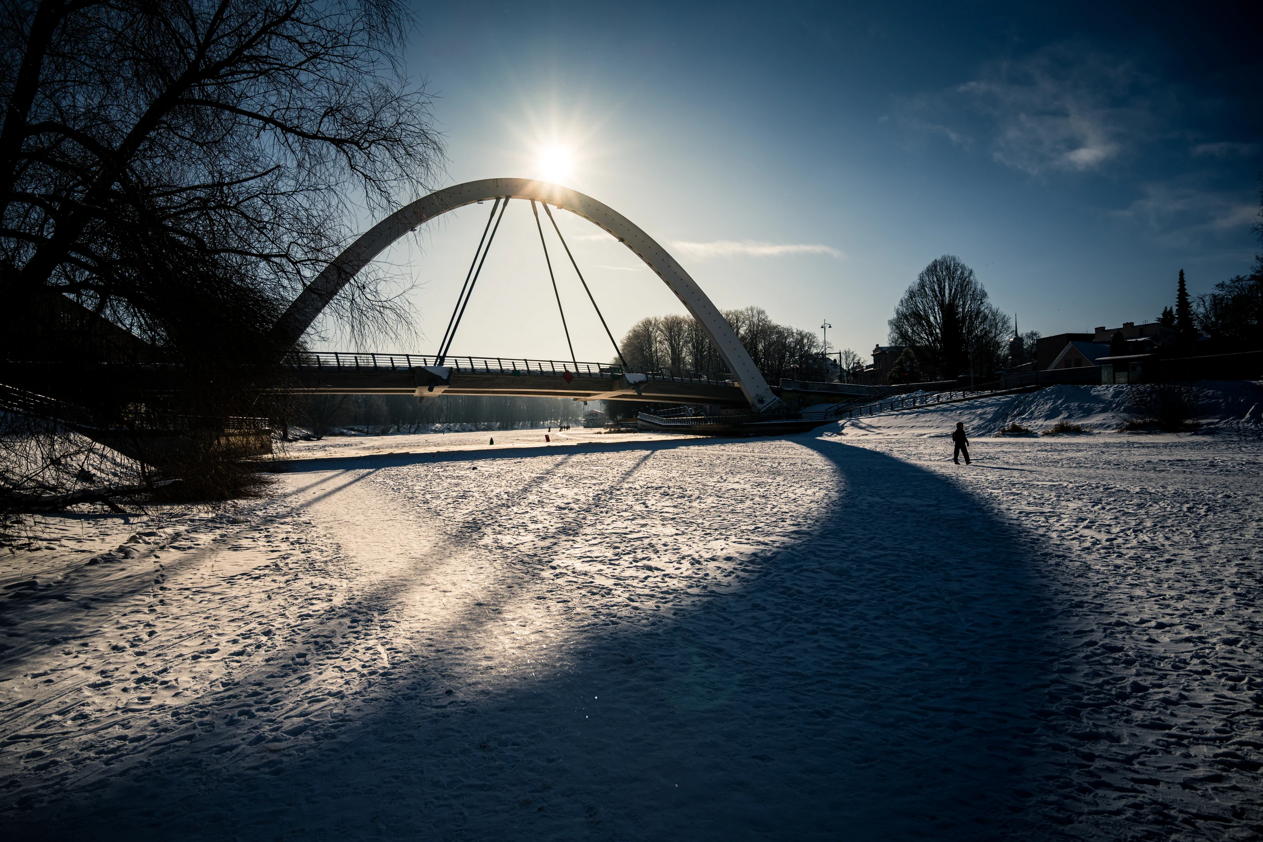 The sun above the arch of Vabadussild, with its curved shadow and a walker on the snow-covered Emajõgi River.